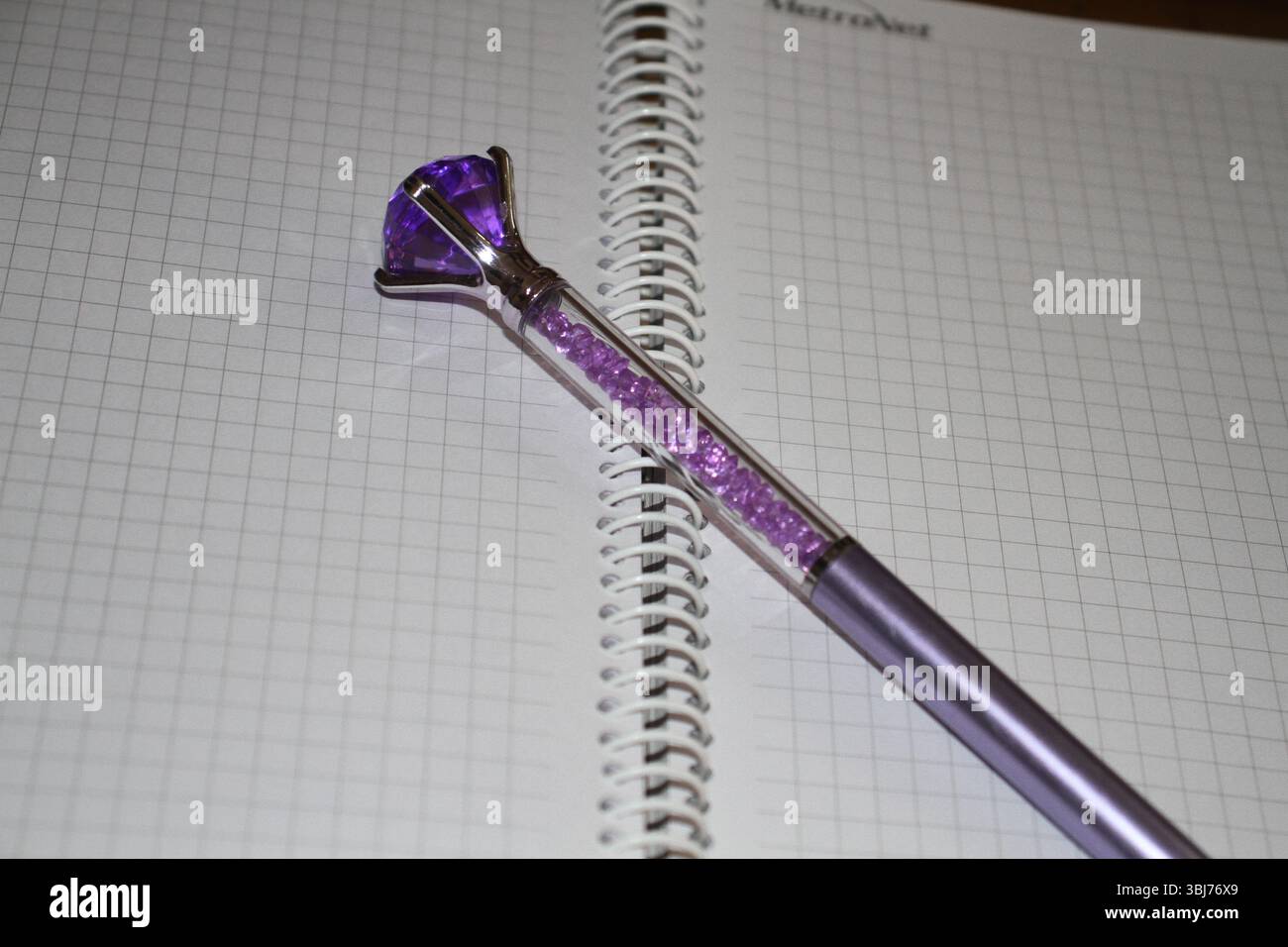 Una penna viola elegante e decorativa posizionata diagonalmente su un notebook grafico aperto con rilegatura a spirale. La penna presenta un grande cristallo simile a una gemma nella parte superiore. Foto Stock