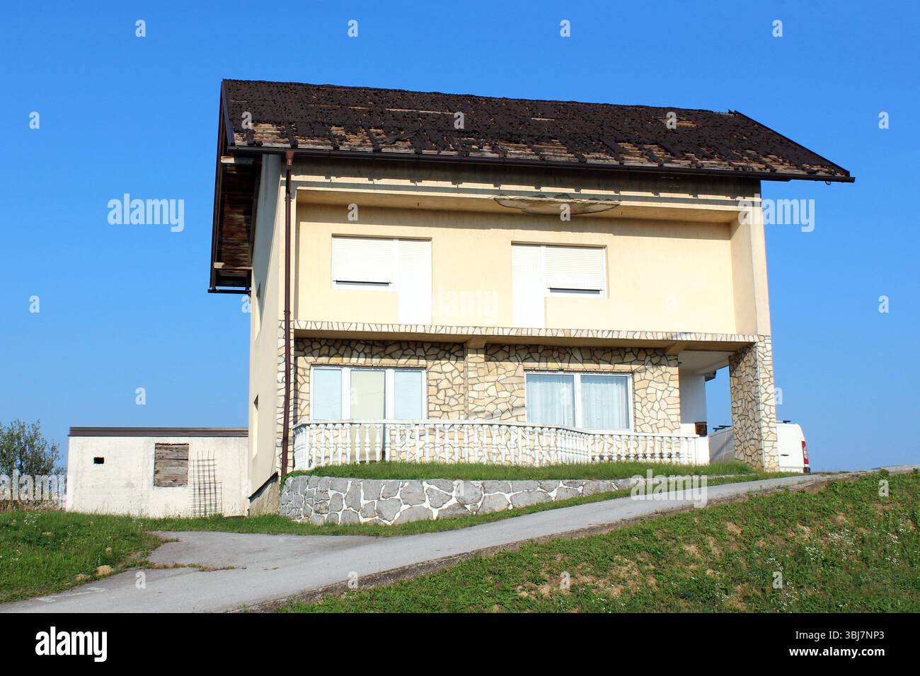 Casa a due piani con facciata in pietra e pareti color crema adagiata su un pendio erboso, tetto visibilmente danneggiato e piastrelle mancanti, balcone bianco e persiane chiuse Foto Stock