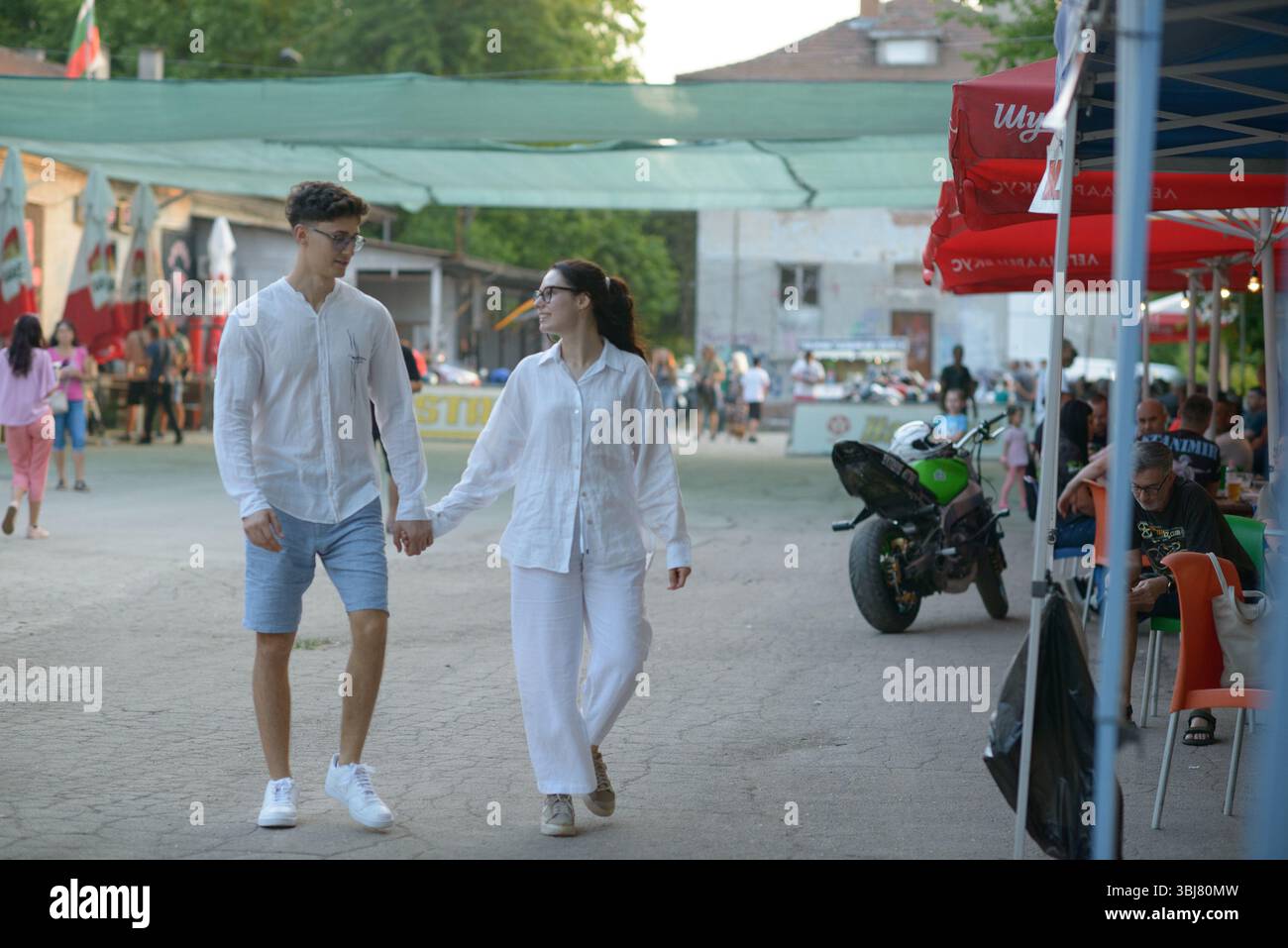 Una giovane coppia cammina mano nella mano durante la prima serata del Moto Rock Fest 2025 a Ruse, Bulgaria, il 13 giugno. Vestiti con abiti bianchi coordinati, passeggiano per il luogo dell'evento mentre motociclisti, famiglie e frequentatori del festival si godono l'atmosfera vivace sotto gli ombrelloni rossi e il panno verde. Una moto sportiva verde è parcheggiata nelle vicinanze e la gente del posto è seduta ai tavoli sulla destra. Il festival internazionale annuale attrae appassionati di motociclismo e rock da tutta la Bulgaria, la Romania e la Macedonia del Nord, trasformando la città in un centro di musica, cultura motoria e celebrità della comunità Foto Stock