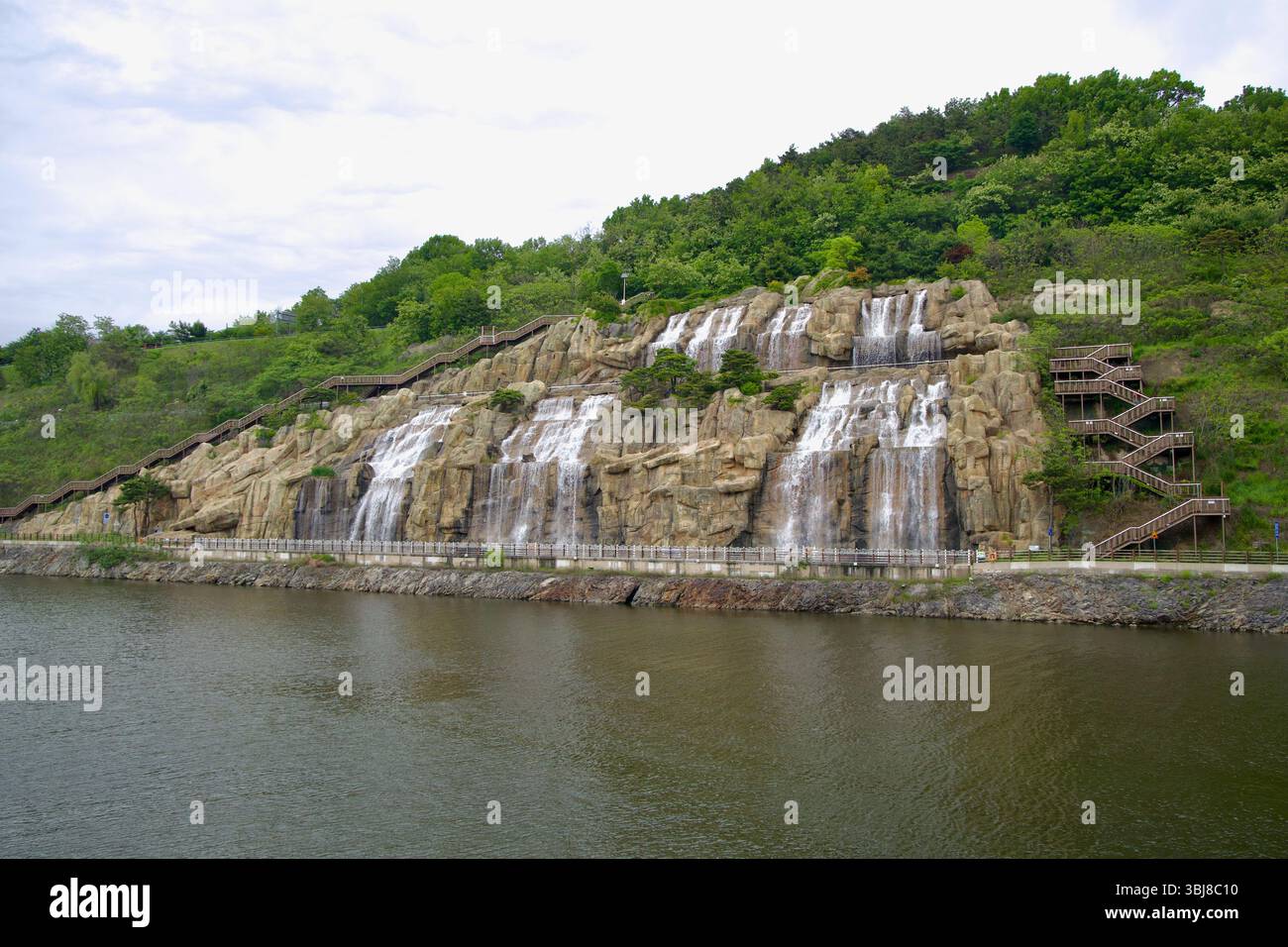 Incheon, Corea del Sud - 19 maggio 2025: Le cascate Ara riversano su terrazze di roccia scolpite nel canale Ara Waterway, incorniciate da scale di legno zigzagate e lussureggianti S. Foto Stock