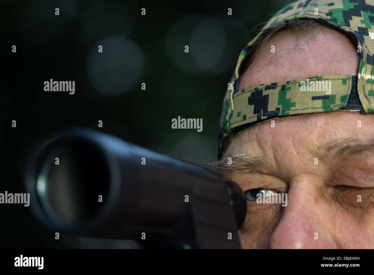 Primo piano di un uomo con un cappuccio mimetico che punta una pistola. Concetto di caccia, tiro o addestramento militare Foto Stock