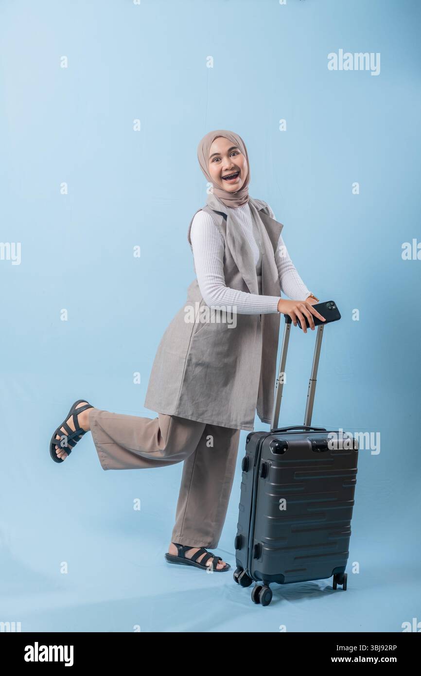 Una donna che indossa un gilet grigio e pantaloni tan sorride e tiene in mano una valigia. La valigia è nera e dispone di ruote Foto Stock
