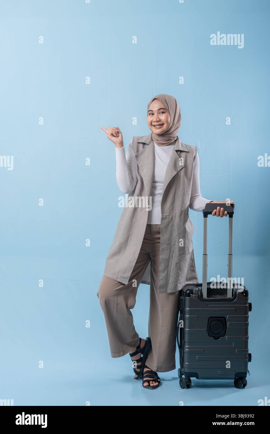 Una donna che indossa una camicia bianca e un gilet grigio tiene in mano una valigia e indica qualcosa di importante Foto Stock