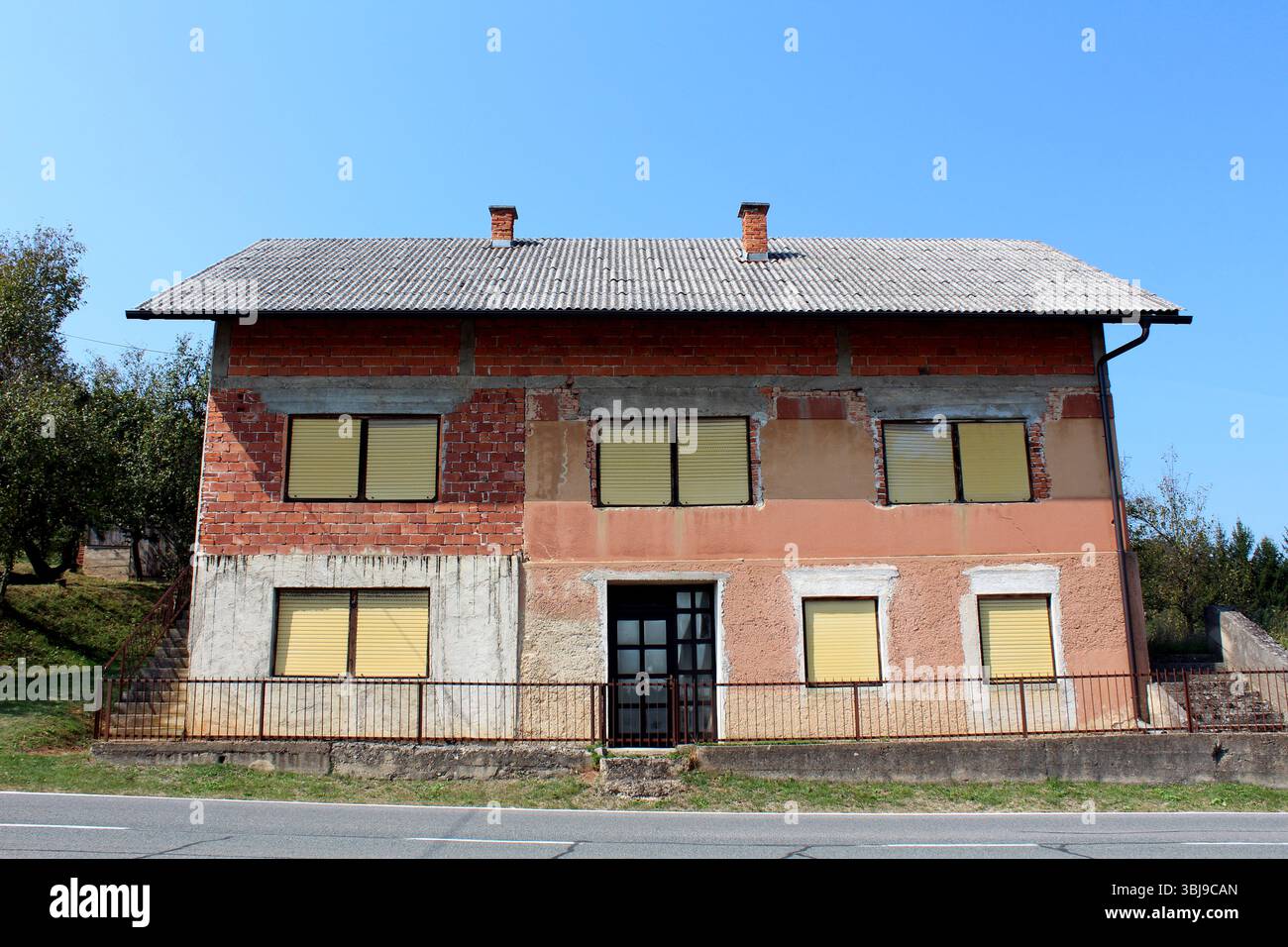 Casa in mattoni incompiuta a due piani con persiane gialle e dettagli in cemento si erge accanto a una strada, mostrando muratura esposta e un cortile inclinato con alberi Foto Stock