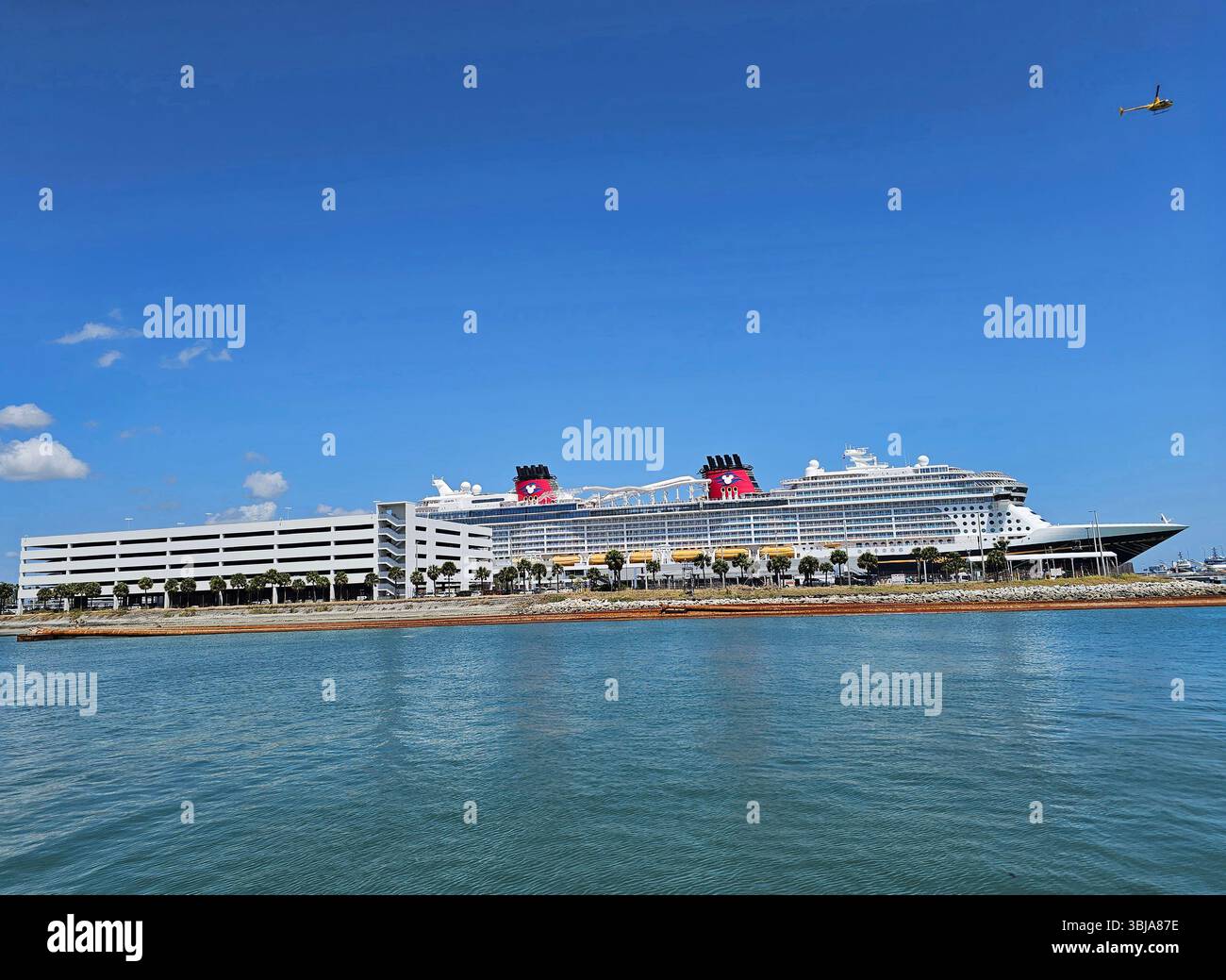 Nave da crociera Disney a Port Canaveral, Florida. La nave è più alta del parcheggio a 6 piani. Un elicottero giallo brillante vola sopra la testa. Foto Stock