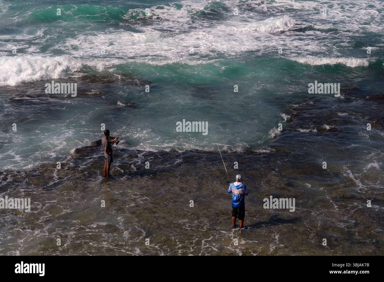 Guardando dall'alto verso il basso due uomini che pescano dalle rocce con i piedi in acqua sulla riva di una spiaggia sulla costa di Durban, in Sudafrica. Foto Stock