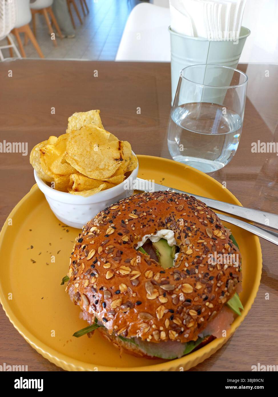 Sandwich con bagel multigrano con strati di prosciutto avocado e verdure servito con patatine croccanti su un piatto giallo accanto a un bicchiere d'acqua Foto Stock