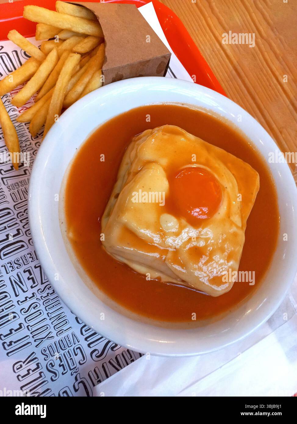 Primo piano di un tradizionale panino francesinha portoghese ricoperto di formaggio fuso e di un uovo fritto, servito in una ricca salsa a base di birra in una ciotola bianca, Foto Stock