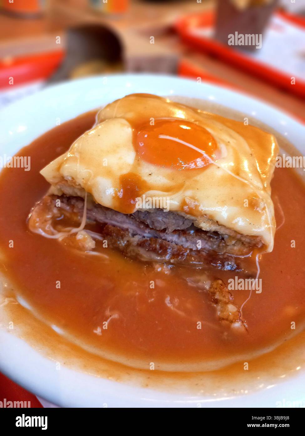 Primo piano di un tradizionale panino francesinha portoghese ricoperto di formaggio fuso e di un uovo fritto, servito in una ricca salsa a base di birra in una ciotola bianca, Foto Stock