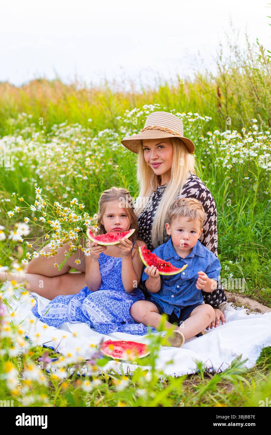 mosca, russia, 30.07.2020 Una donna e due bambini siedono su una coperta in un campo. La donna ha lunghi capelli biondi e indossa un cappello di paglia. I bambini lo sono Foto Stock