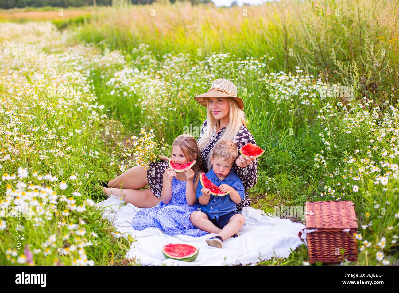 mosca, russia, 30.07.2020 Una donna e due bambini si godono l'anguria in un campo di fiori selvatici. La donna ha lunghi capelli biondi e indossa un cappello di paglia. Foto Stock