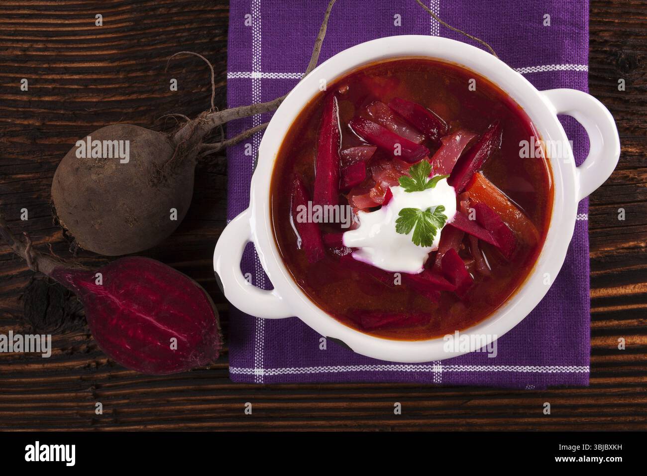 Deliziosa zuppa borsch ucraina con verdure fresche di barbabietola su un panno viola su un tavolo rustico in legno, vista dall'alto. Cucina tradizionale europea Foto Stock