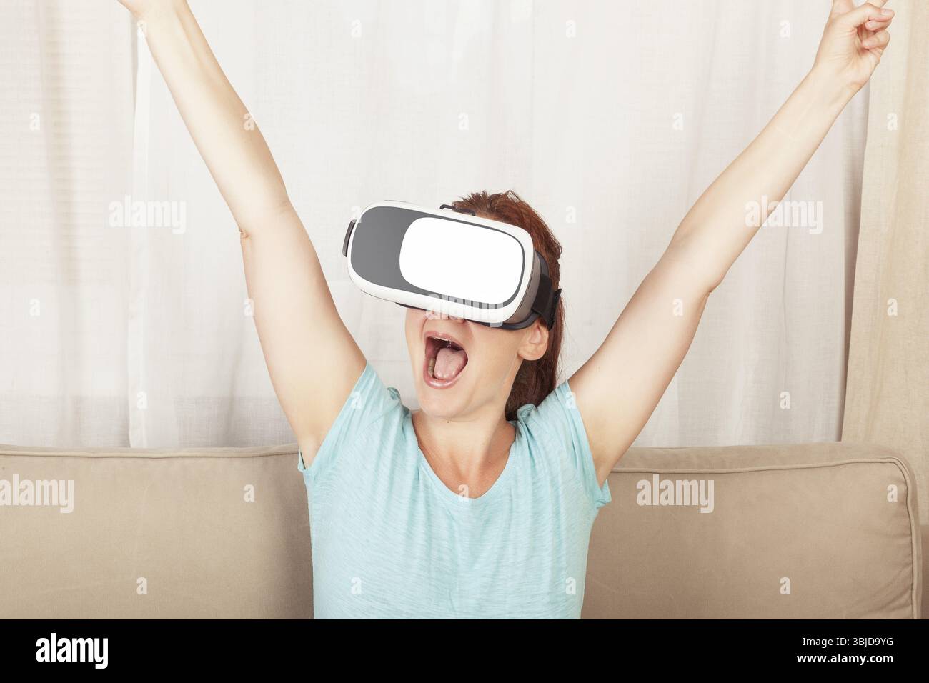 Giovani donne belle vince nella realtà virtuale sul divano in salotto. Videogamer femminile con le mani in alto e guanti VR. Intrattenimento futuro Foto Stock