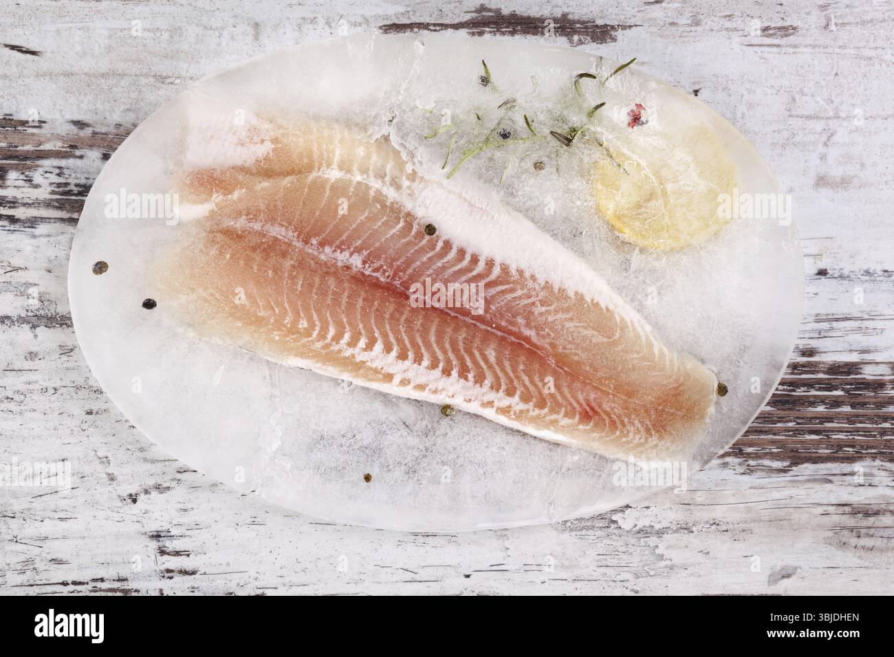Filetto di pesce congelato in ghiaccio con grani di pepe, erbe fresche e limone su fondo bianco testurizzato in legno. Piatti di pesce in stile mediterraneo country Foto Stock