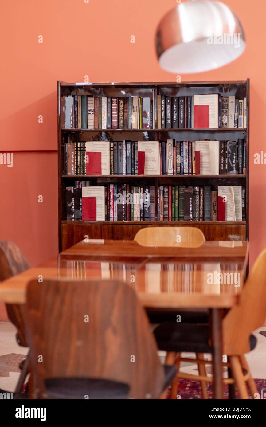 Accogliente angolo di lettura in un interno moderno con un tavolo in legno, sedie colorate e una libreria ben rifornita contro una calda parete arancione Foto Stock