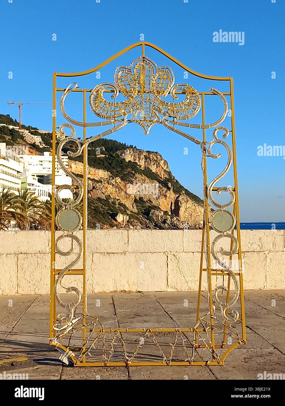 Sesimbra, Portogallo. 20 dicembre 2024 Una cornice ornamentale in metallo dorato che si erge vuota contro le scogliere costiere e lo sfondo azzurro del mare, fiancheggiata da Palm tre Foto Stock
