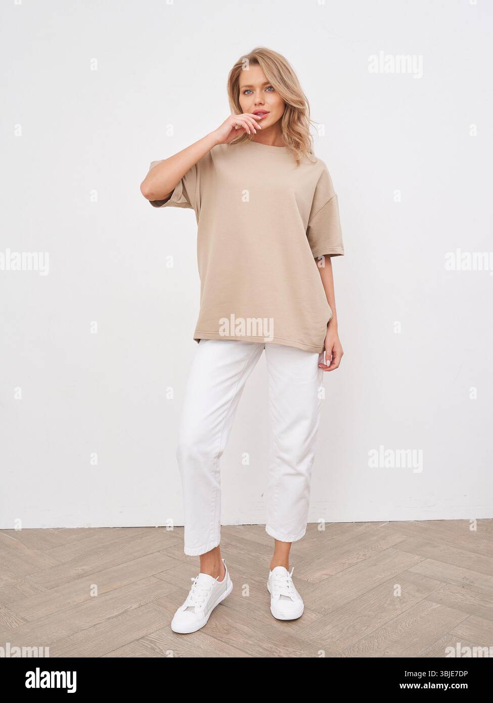 Modello di moda casual con top beige e pantaloni bianchi, in piedi con sicurezza su pavimento in legno. Capelli biondi con onde morbide, espressione neutra Foto Stock