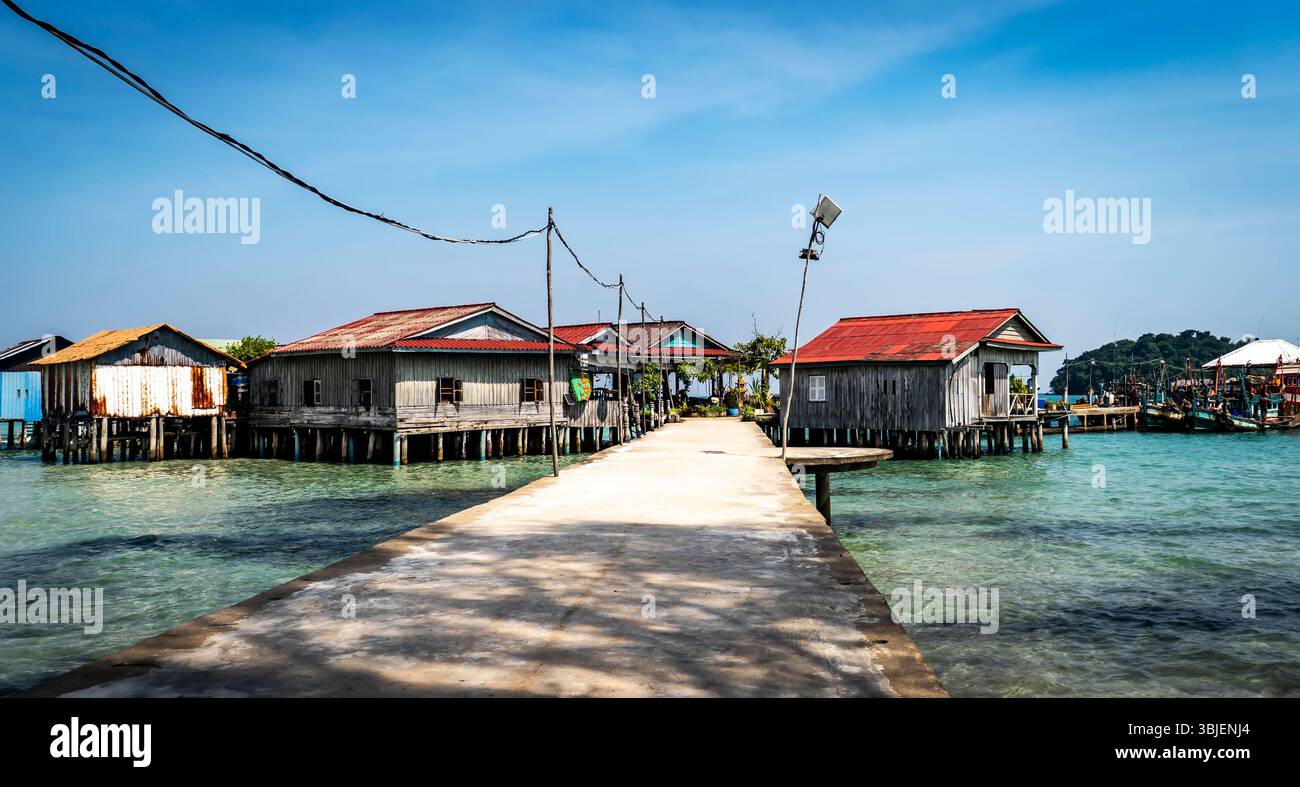 Una vista panoramica di case in legno su palafitte su limpide acque turchesi, collegate da un passaggio pedonale in cemento. Il cielo è blu brillante con qualche nuvola, creato Foto Stock