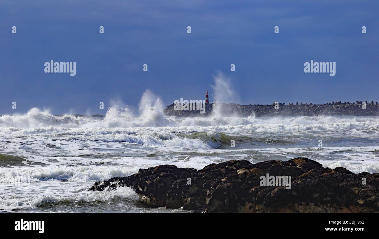Cabedelo, Viana do Castelo, Portogallo - 5 marzo 2024: Le onde si infrangono drammaticamente contro la frangia con un faro alto sul retro Foto Stock