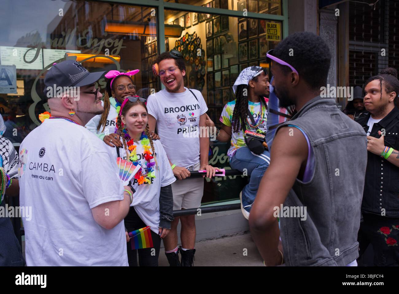 Un gruppo di persone sorridenti, molte delle quali indossano abiti e accessori a tema arcobaleno, si trova all'esterno di un edificio. Molti indossano le camicie Brooklyn Pride. Foto Stock