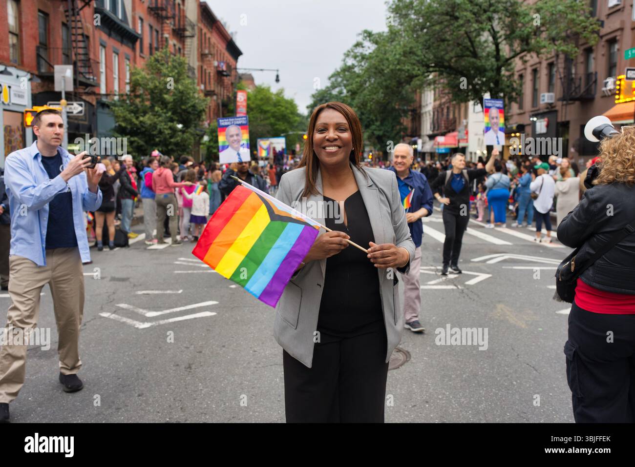 Una donna sorride mentre tiene in mano una bandiera arcobaleno a una parata del Pride. Indossa un blazer grigio e pantaloni neri. Anche le persone sullo sfondo sono parti Foto Stock