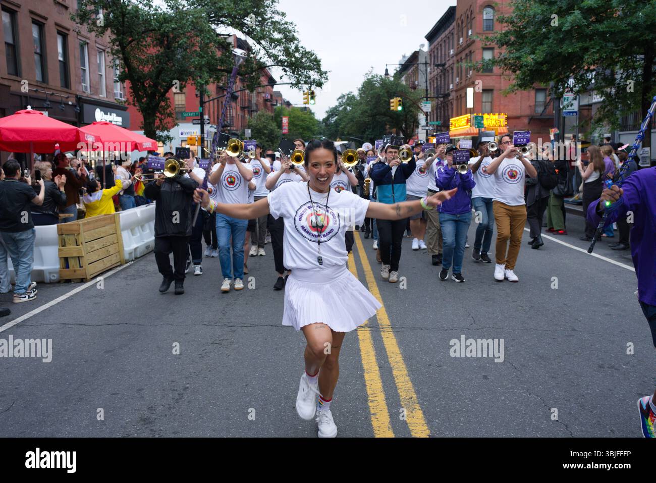 Una donna con una gonna bianca e una camicia conduce una banda lungo una strada di città. La fascia indossa camicie bianche con loghi viola e suona uno strumento in ottone Foto Stock