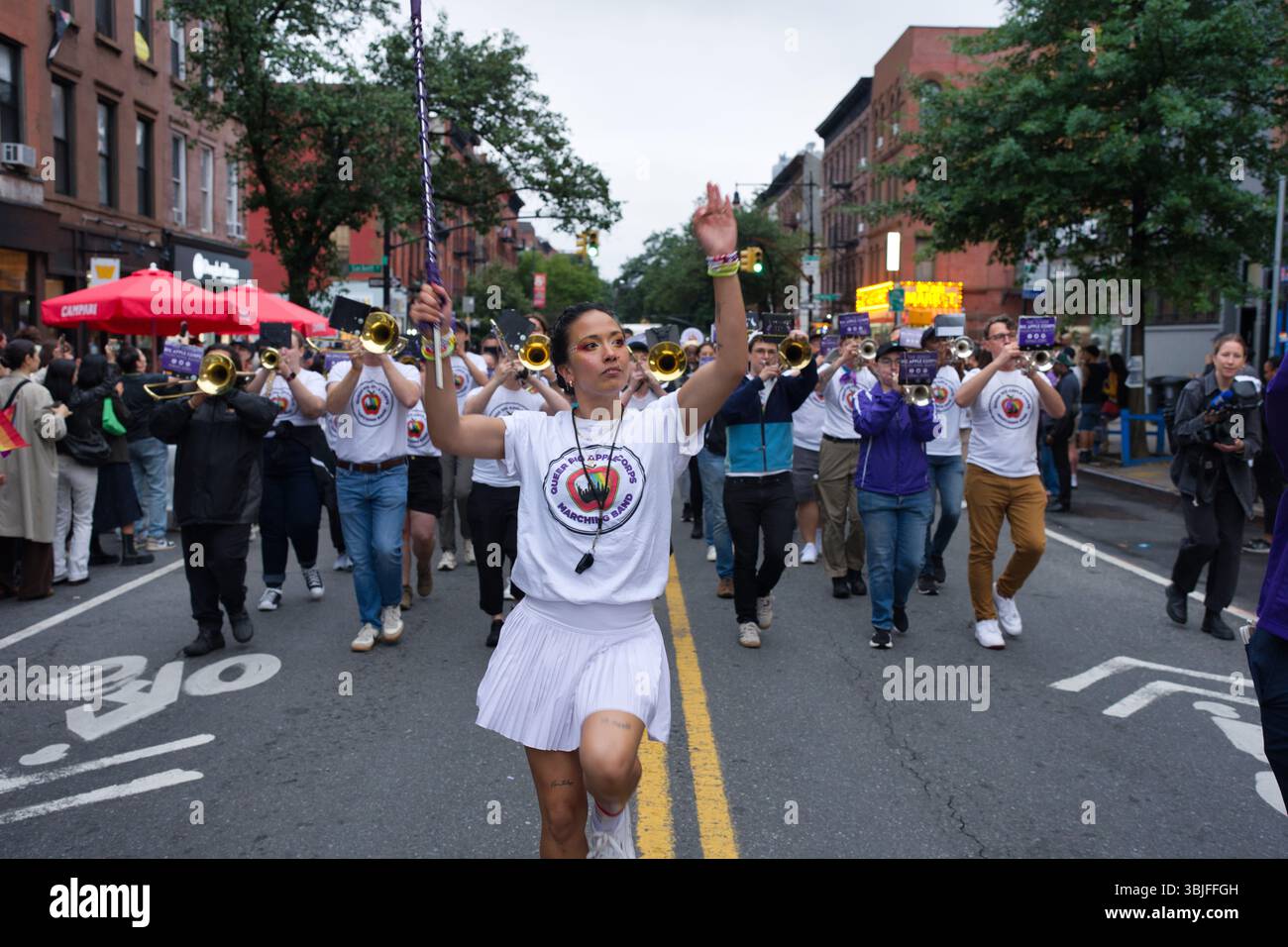 Una donna guida una banda lungo una strada cittadina. Indossa una gonna bianca e una maglietta con il logo della fascia e tiene in mano una bacchetta. I membri della band indossano il bianco Foto Stock