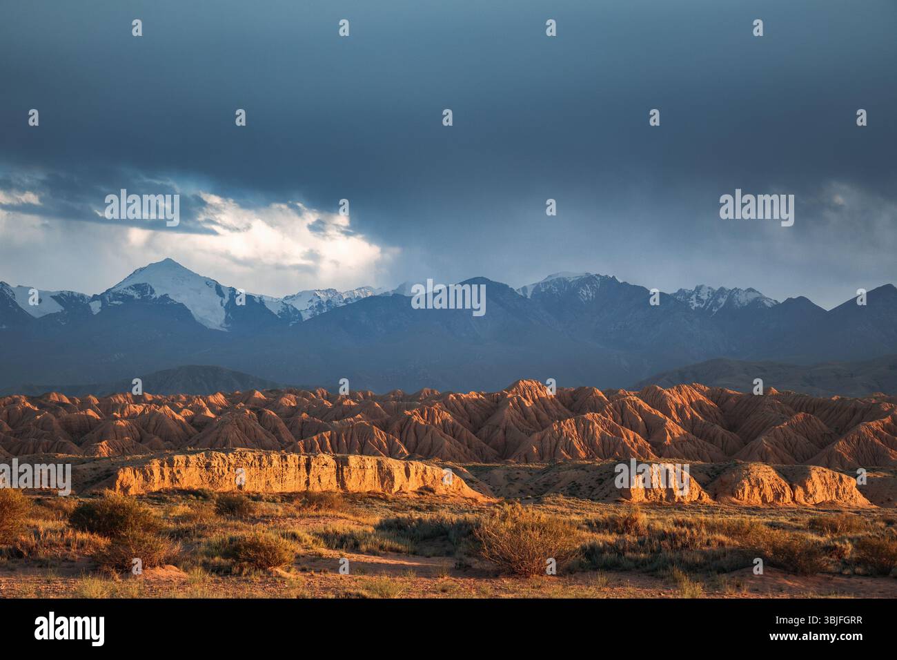 Le accattivanti formazioni rocciose rosse scolpite del Fairytale Canyon illuminate dal sole al tramonto, con le montagne innevate di Tian Shan sulla schiena Foto Stock