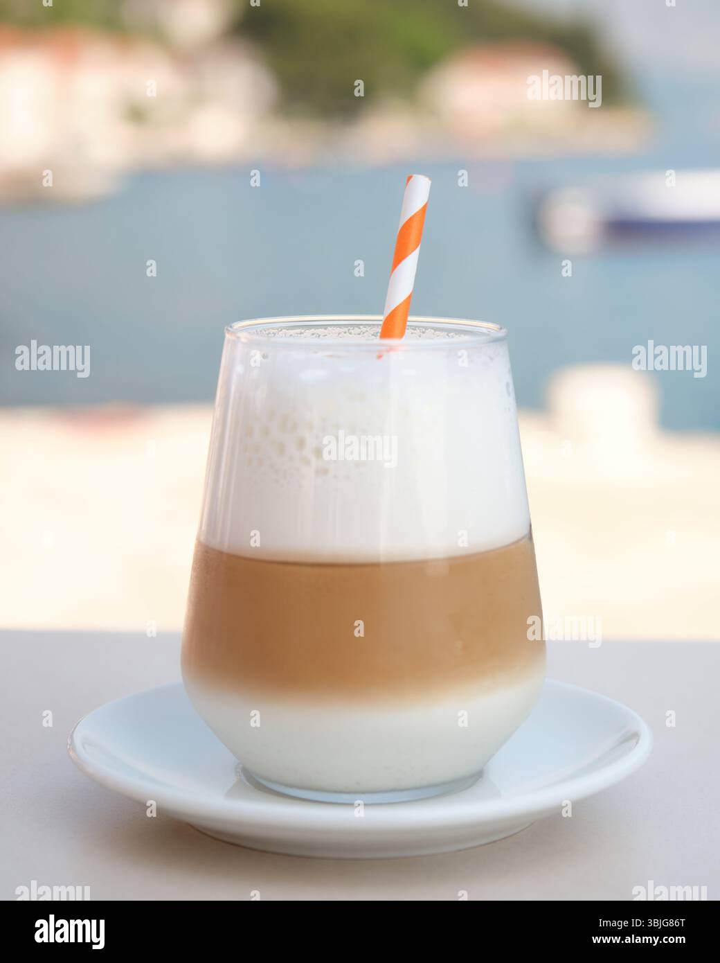 Latte ghiacciato con schiuma di latte e paglia a righe arancioni fotografato su un tavolino con un morbido sfondo costiero Foto Stock