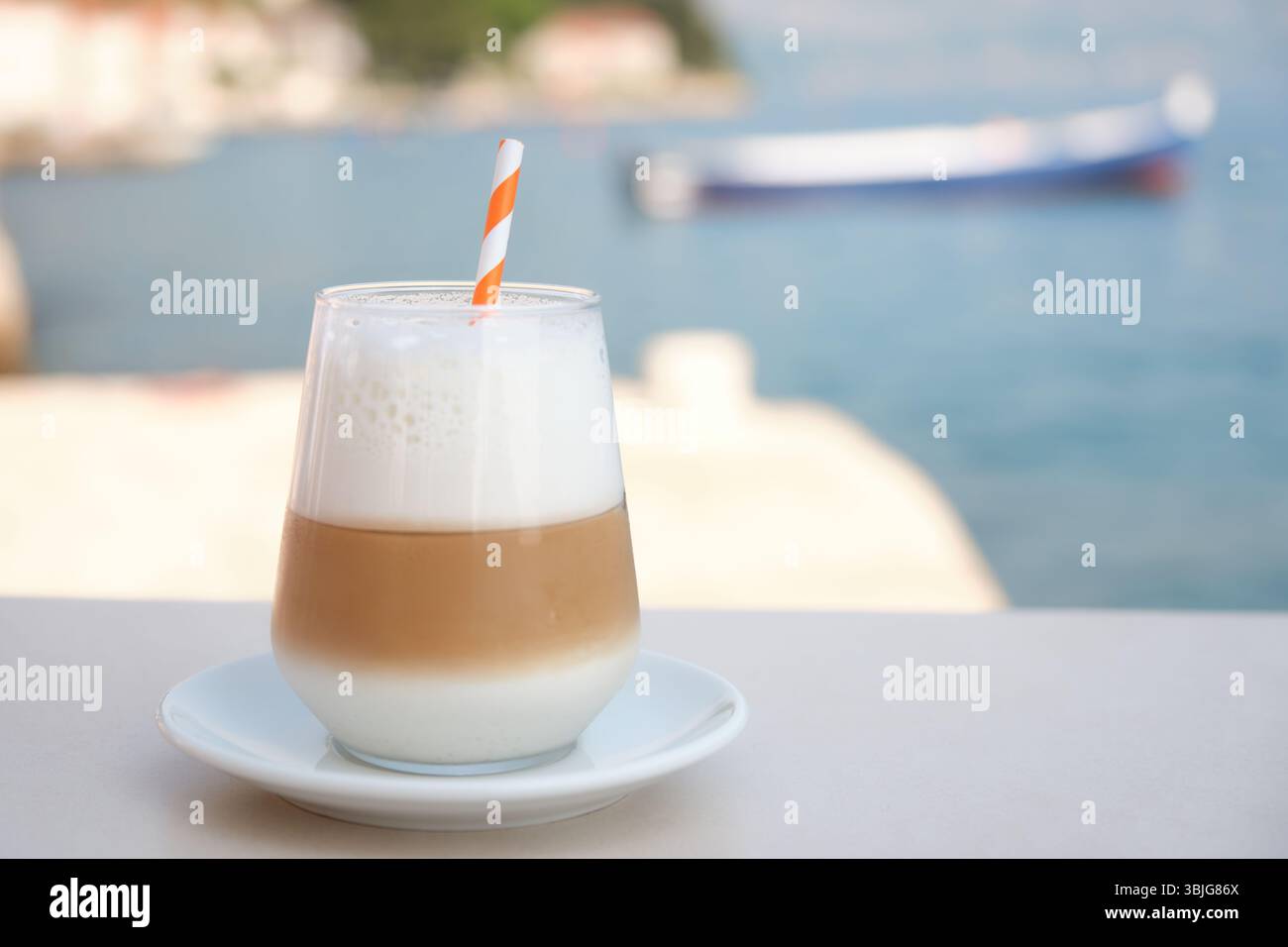 Bicchiere di latte ghiacciato con schiuma di latte e paglia su un tavolo vicino al mare. Barca sfocata ed edifici costieri sullo sfondo Foto Stock