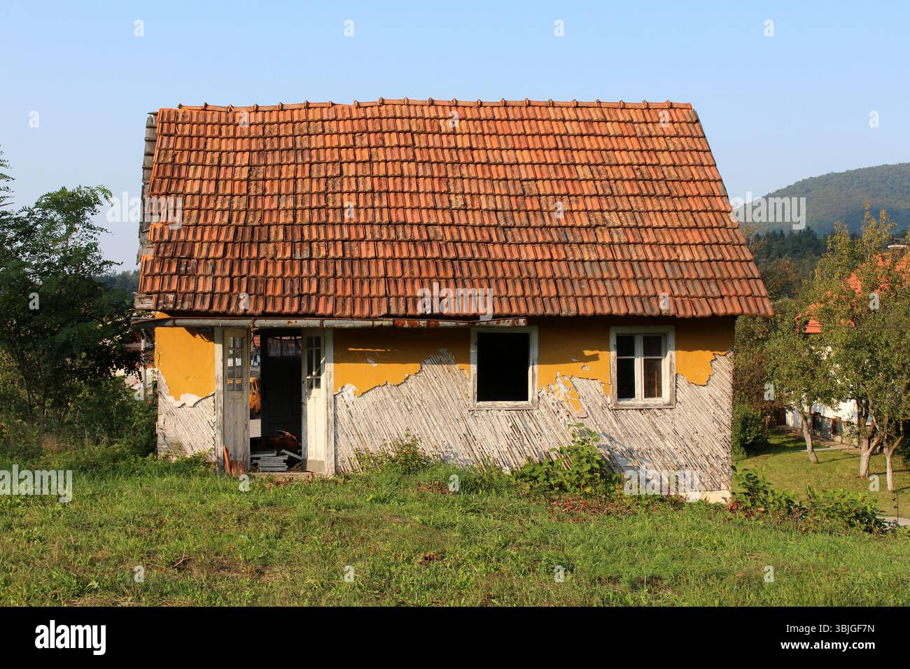 Una piccola casa rurale abbandonata con un tetto in piastrelle rosse intemprate e vernice gialla sbucciante si erge in un campo erboso, le sue porte e finestre si aprono sull'elem Foto Stock