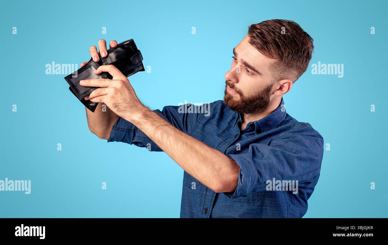 Fallimento, povertà e disoccupazione. Miserabile giovane uomo che guarda nel portafoglio vuoto senza soldi su sfondo blu Foto Stock