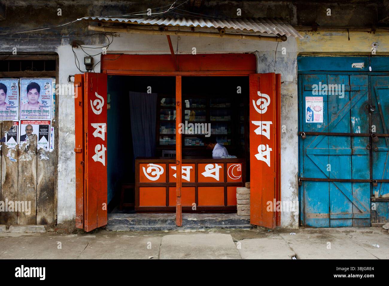 Piccolo negozio di medicine a Jessore, Bangladesh, con cartellonistica bengalese, persiane arancioni e un uomo seduto dietro un bancone di prodotti farmaceutici confezionati. Foto Stock