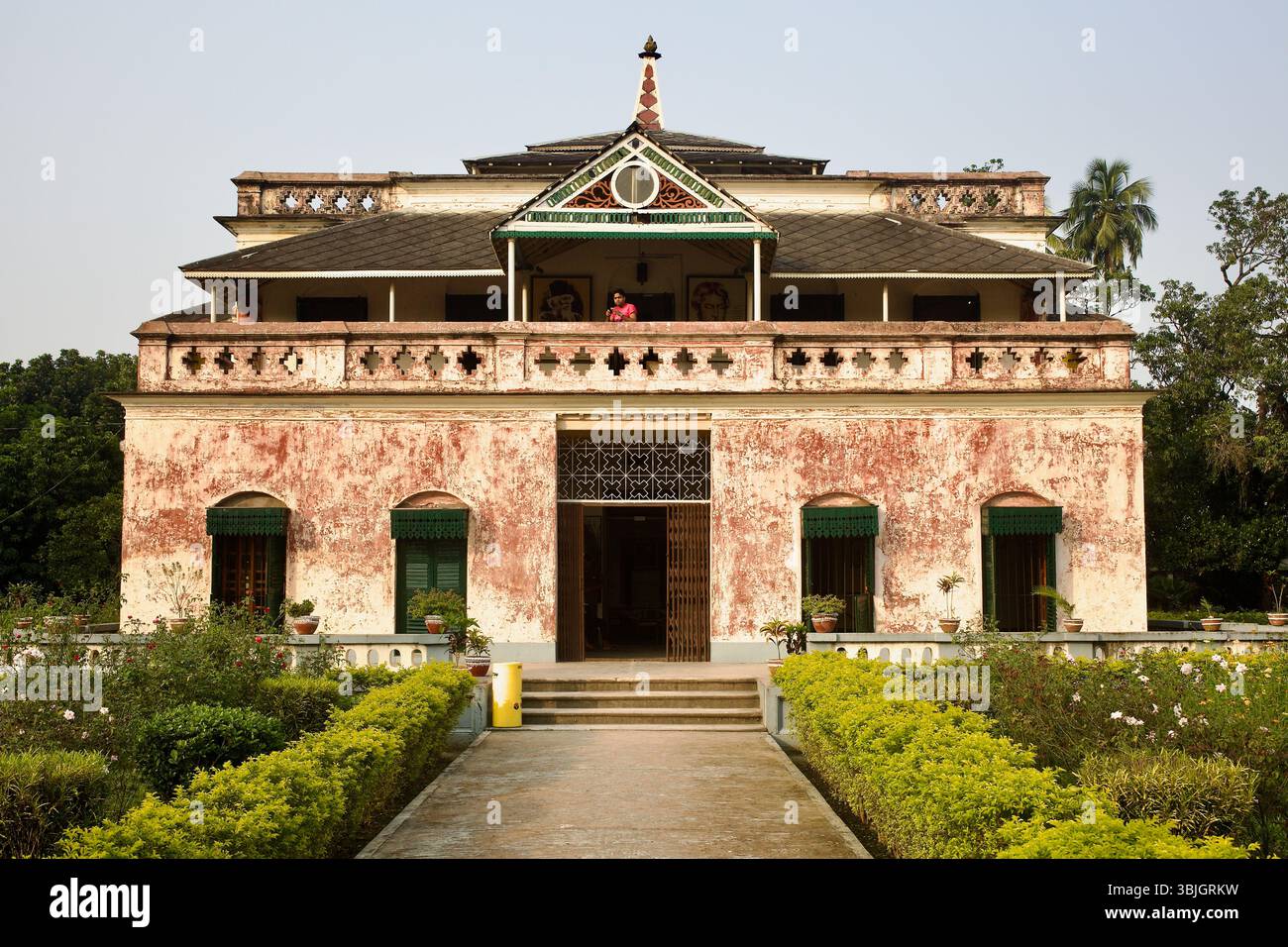 La facciata centrale di Shilaidaha Kuthibari, con le sue pareti in patina rosa, il balcone ad arco e i dettagli di epoca coloniale, visti sotto la luce soffusa del pomeriggio. Foto Stock