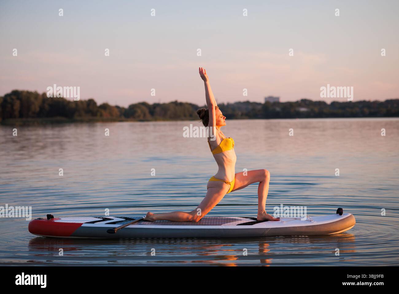 Donna in forma che fa yoga sul paddleboard all'alba, tenendo in posa il guerriero con equilibrio e grazia, circondata da acqua tranquilla e luce naturale soffusa. Foto Stock