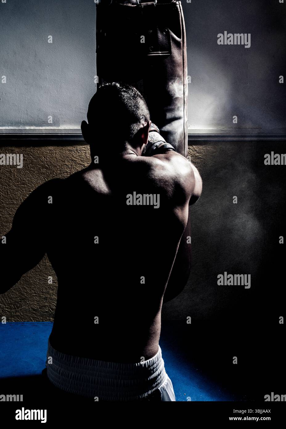 Un pugile maschile, visto dalla schiena e indossato pantaloncini da boxe bianchi, lancia un pugno potente a una borsa pesante in una palestra poco illuminata. Foto Stock
