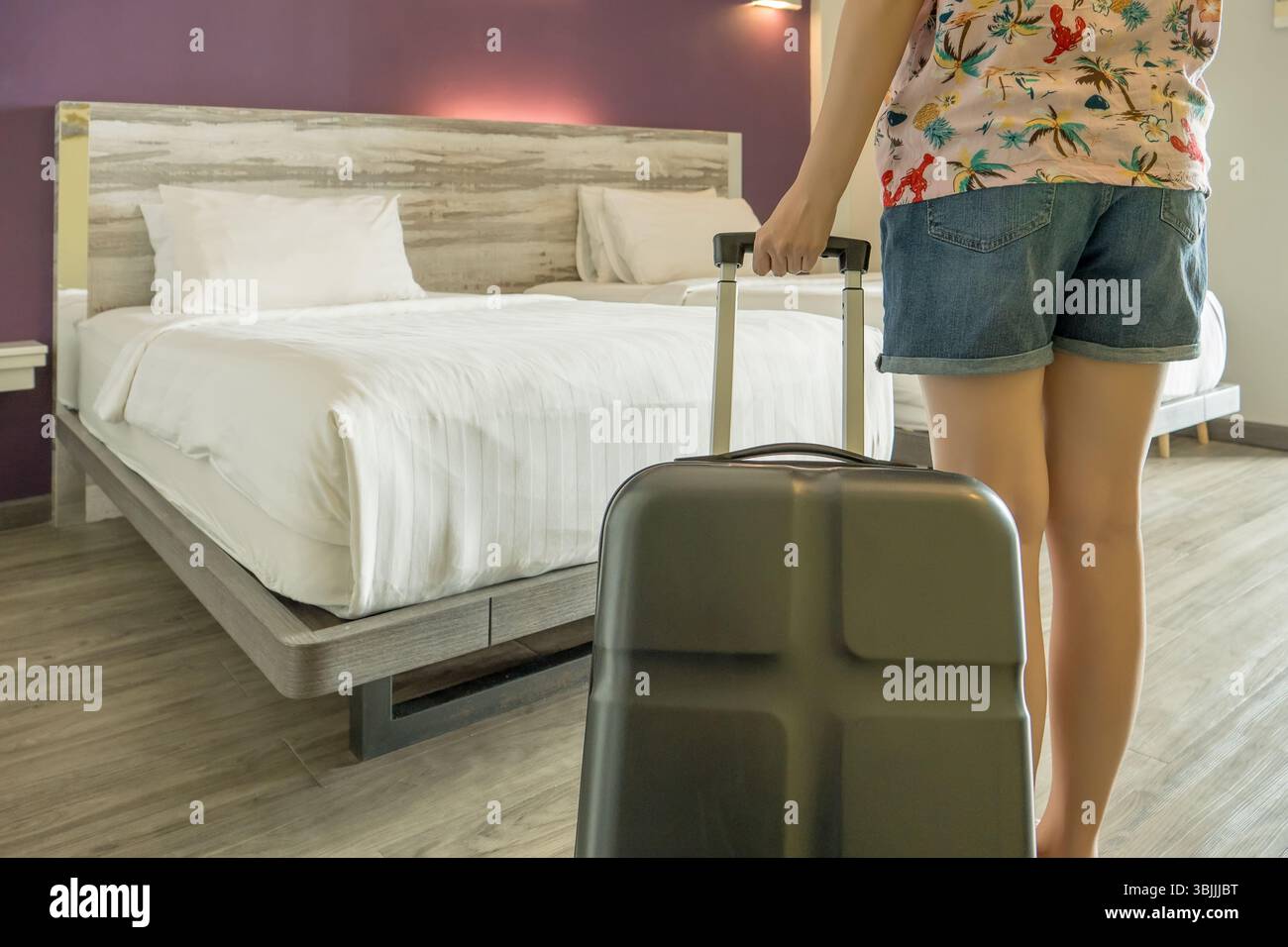 vista sul retro del turista in abbigliamento informale. arrivo in camera con trolley dopo il check-in, letto pulito con lenzuola bianche sullo sfondo Foto Stock