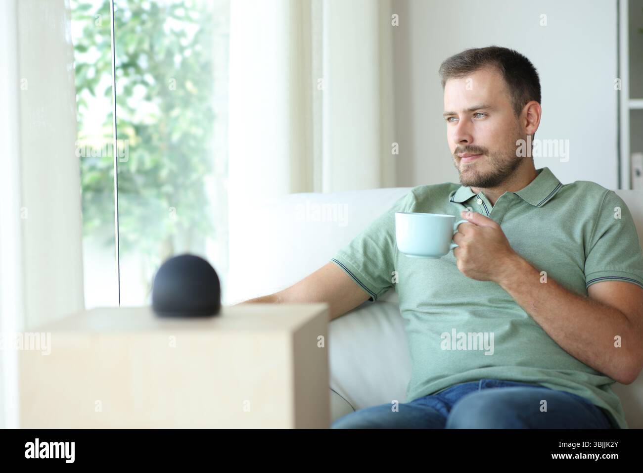 Uomo rilassato con altoparlante intelligente che beve caffè seduto sul divano di casa Foto Stock
