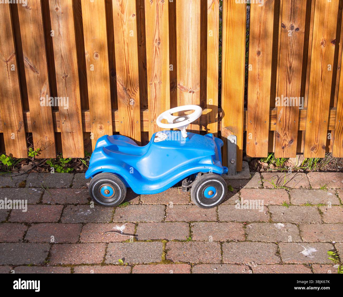 Un'auto giocattolo blu parcheggiata accanto a una recinzione di legno, adagiata su un sentiero in mattoni. La vettura presenta un design semplice con volante ed è posizionata al sole Foto Stock