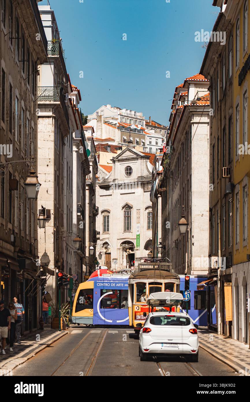 Il classico tram giallo e il moderno tram attraversano una strada stretta e soleggiata costeggiata da vecchi edifici a Lisbona, Portogallo - 11 giugno 2025 Foto Stock