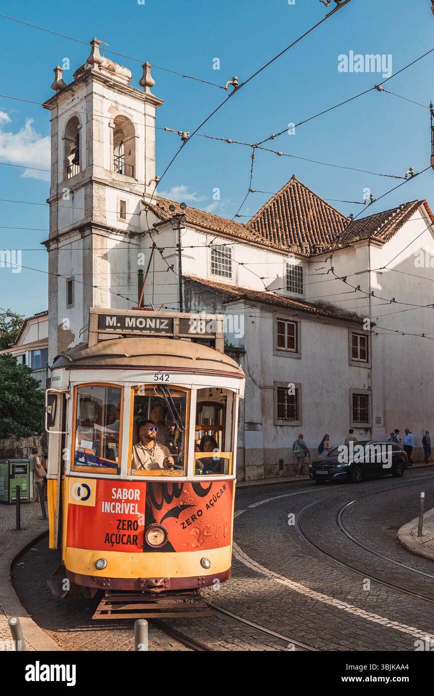 L'iconico tram giallo 28 viaggia attraverso un'affascinante strada di Lisbona, passando davanti a una chiesa storica sotto i cieli soleggiati. Lisbona, Portogallo - 11 giugno 2025 Foto Stock