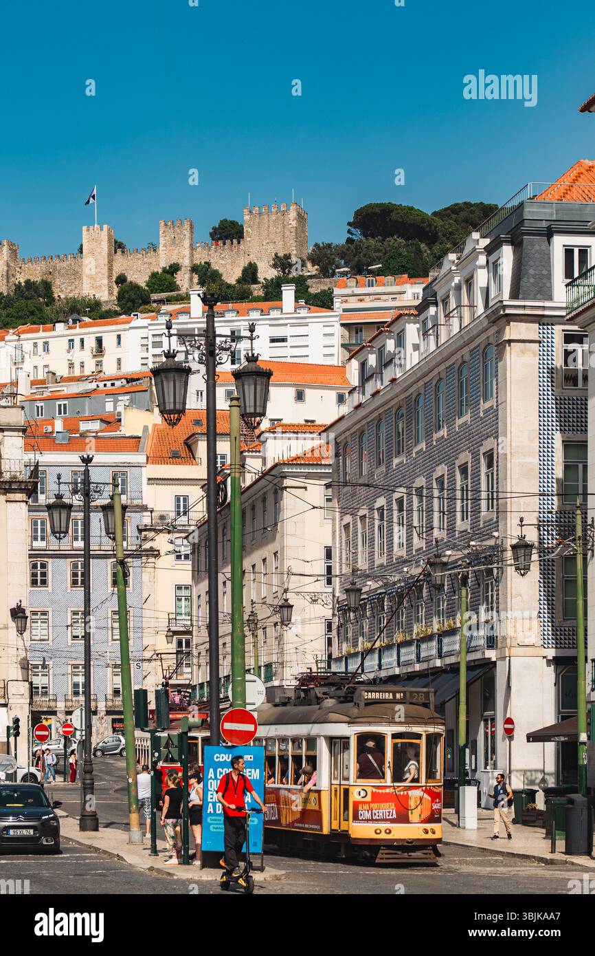 Il classico tram giallo viaggia attraverso le vivaci strade di Lisbona con il castello di Sao Jorge sullo sfondo, catturando la vita e il patrimonio culturale della città. Lisbona, Portogallo - 11 giugno 2025 Foto Stock