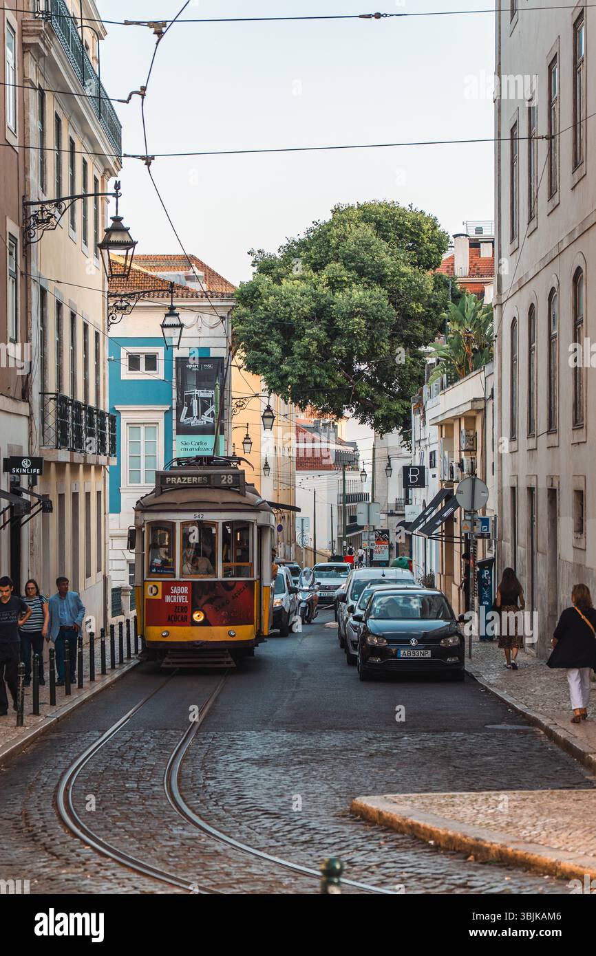 L'iconico tram giallo 28 viaggia lungo un'affascinante strada acciottolata costeggiata da edifici colorati e gente del posto a Lisbona, Portogallo - 11 giugno 2025 Foto Stock