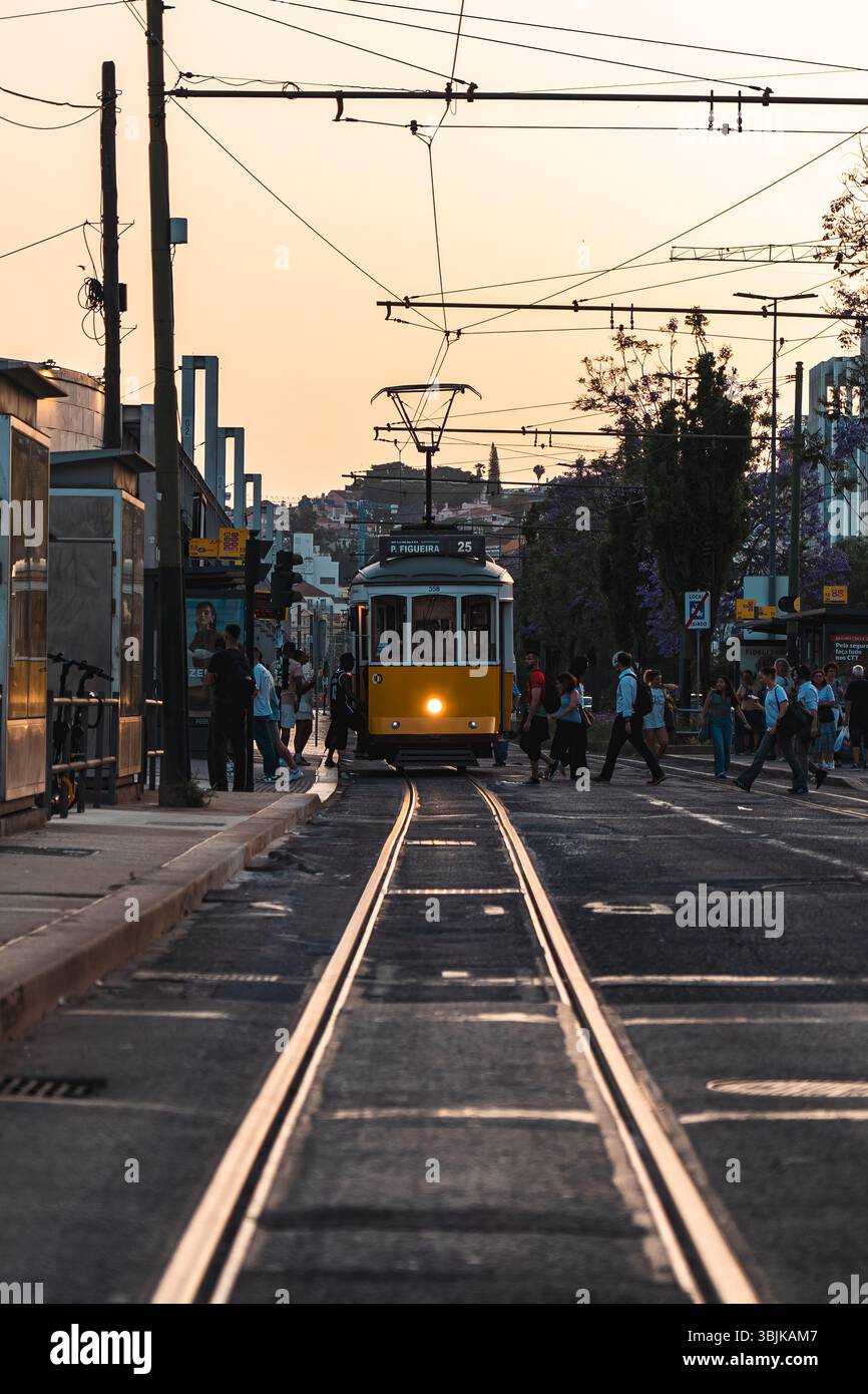 L'iconico tram giallo viaggia attraverso una trafficata strada di Lisbona mentre le persone attraversano al tramonto, catturando la vita urbana quotidiana. Lisbona, Portogallo - 11 giugno 2025 Foto Stock