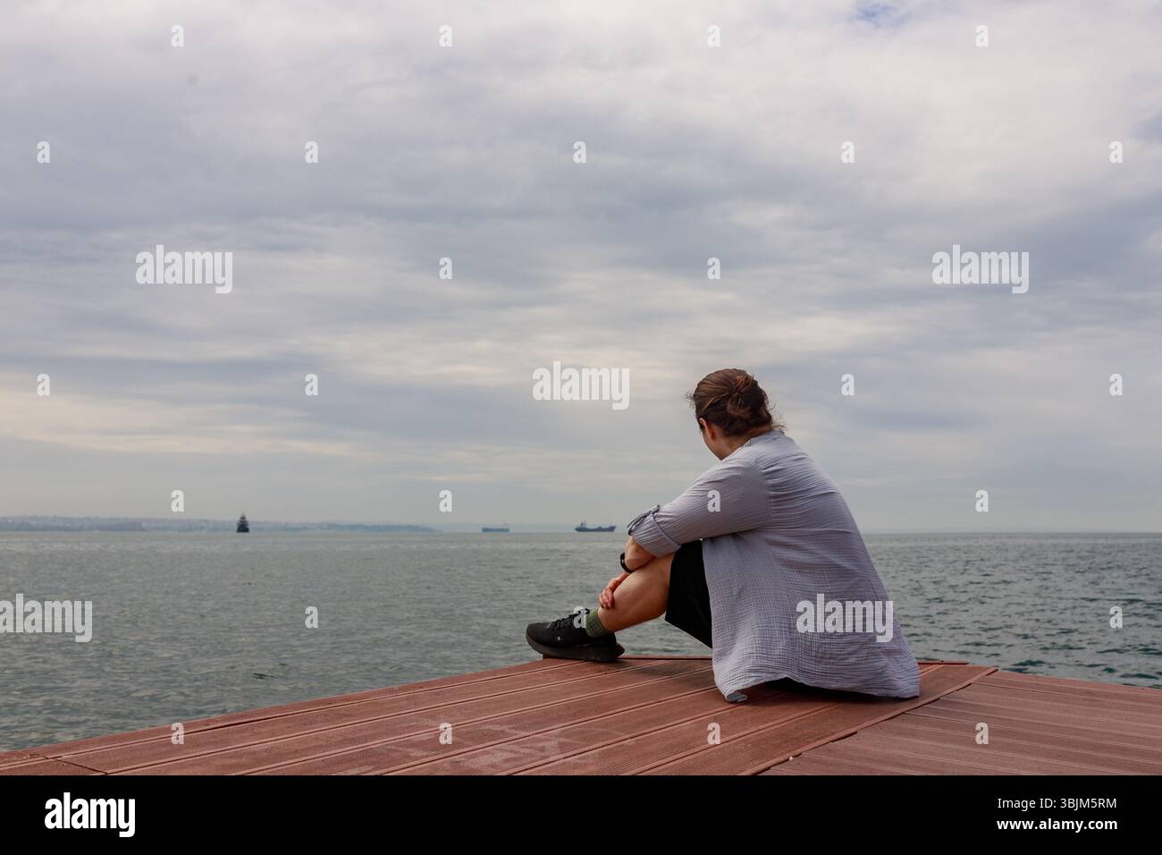 Giovane uomo informale seduto lateralmente sul bordo del molo, guardando verso navi lontane. Foto Stock