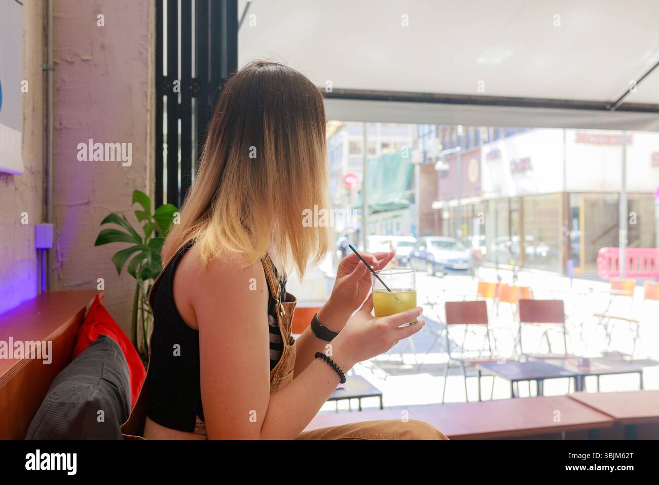 Giovane donna seduta accanto alla finestra con bevanda agli agrumi in mano, caffè interno diurno con sedie all'aperto. Foto Stock