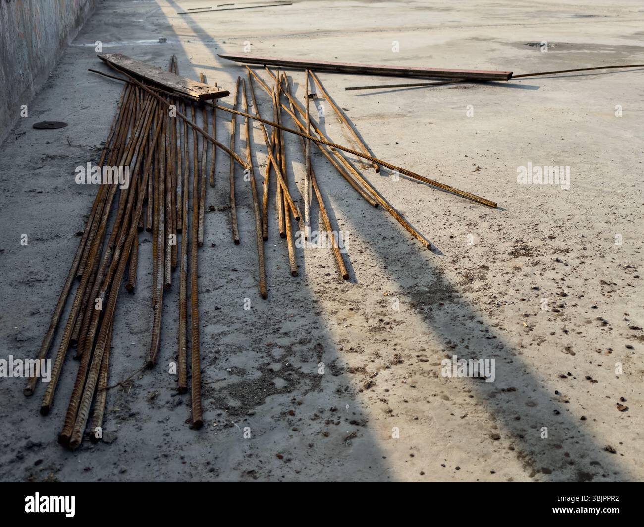 Armature arrugginite e detriti di costruzione sparsi su un terreno di cemento asciutto, riprese in prospettiva con ombre lunghe in condizioni di sole basso. Foto Stock
