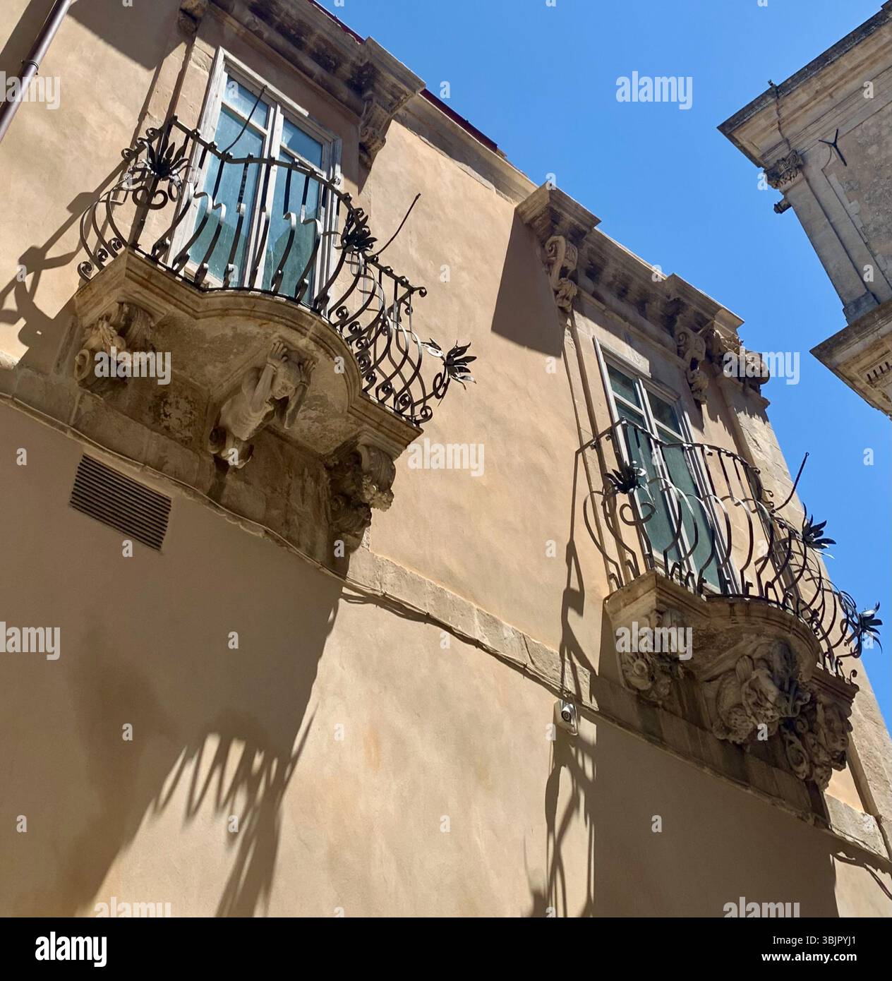 Intricati lavori in pietra barocca sotto un incantevole balcone in ferro battuto a Ragusa, Sicilia, un segno distintivo del patrimonio architettonico della città. Foto Stock
