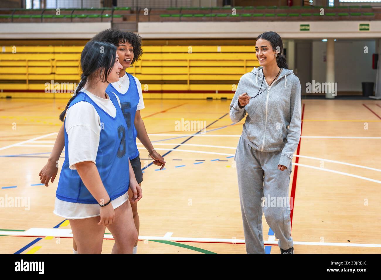 Allenatrice donna con un fischio, che guida i giovani giocatori di basket attraverso esercizi di allenamento in una vibrante atmosfera da palestra piena di energia Foto Stock