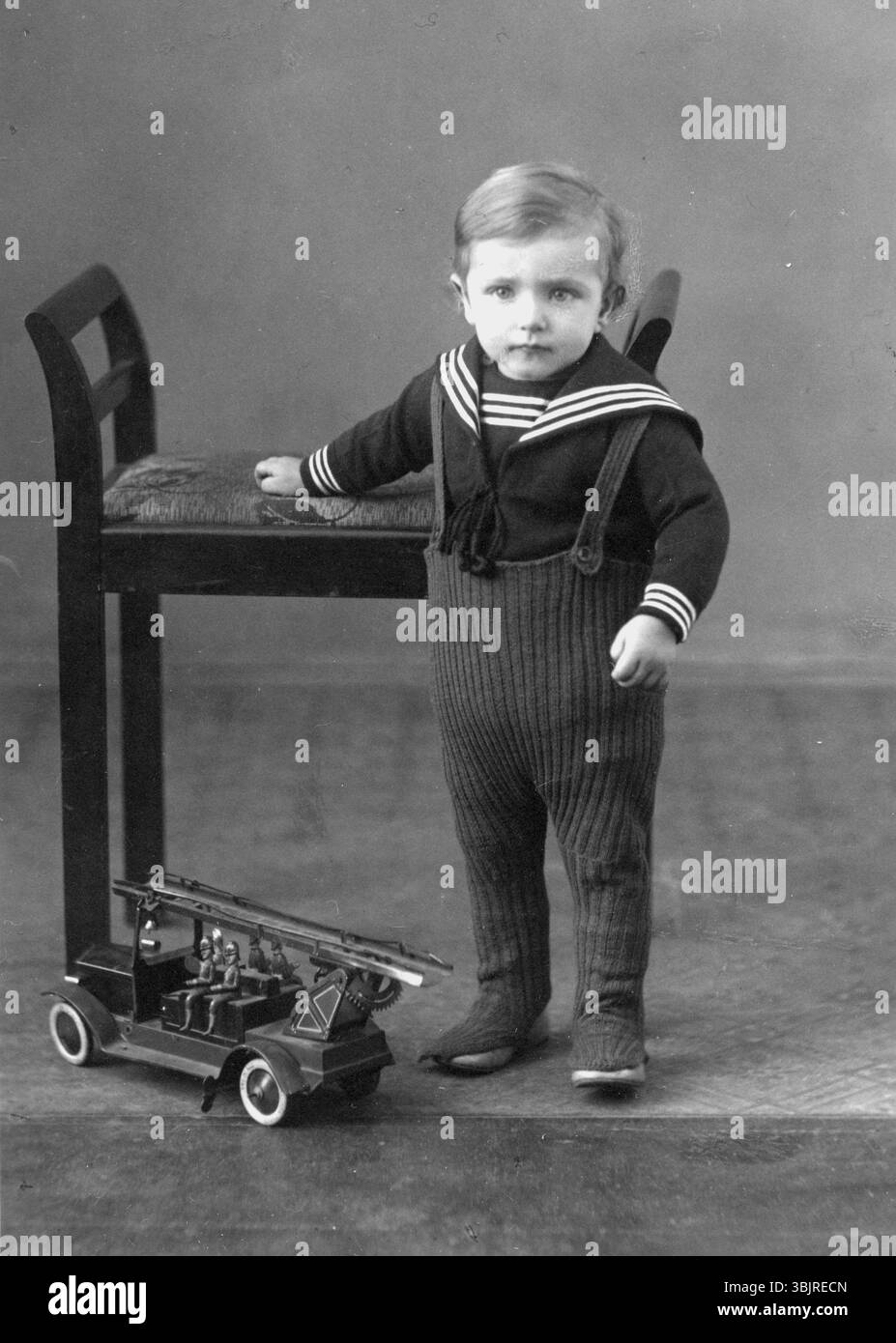 Bambino, giocattolo, motore antincendio, anni '1920, Un bambino in costume da marinaio si trova accanto a una sedia e a un motore antincendio giocattolo, foto storica Foto Stock